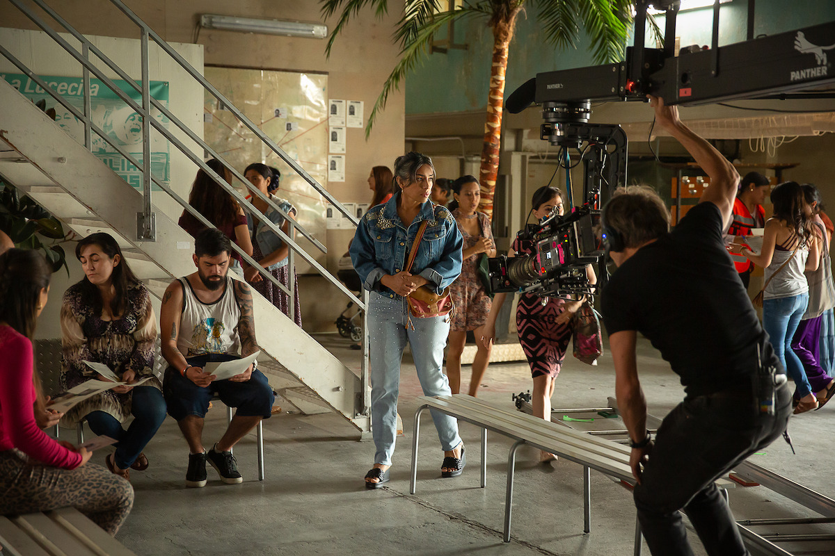 Adriana Paz behind the scenes at La Lucecita. She wears a jean ensemble and clutches her back to her chest.