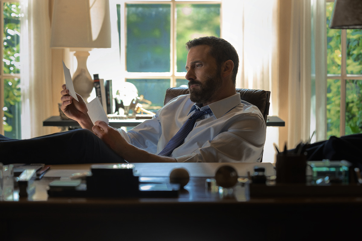 Man in dress shirt and tie sits at a desk in a sunlit office, reading papers with feet up. Desk has pens, papers, and lamps. Large windows and trees outside create a bright, contemplative setting.
