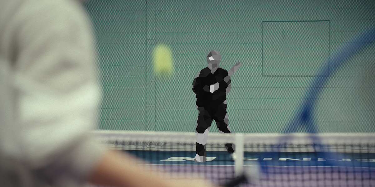 Two people playing tennis indoors; the player in the foreground is preparing to hit the ball while the opponent in the background waits to return the shot. The setting is a gym with a green wall and a net in the middle.