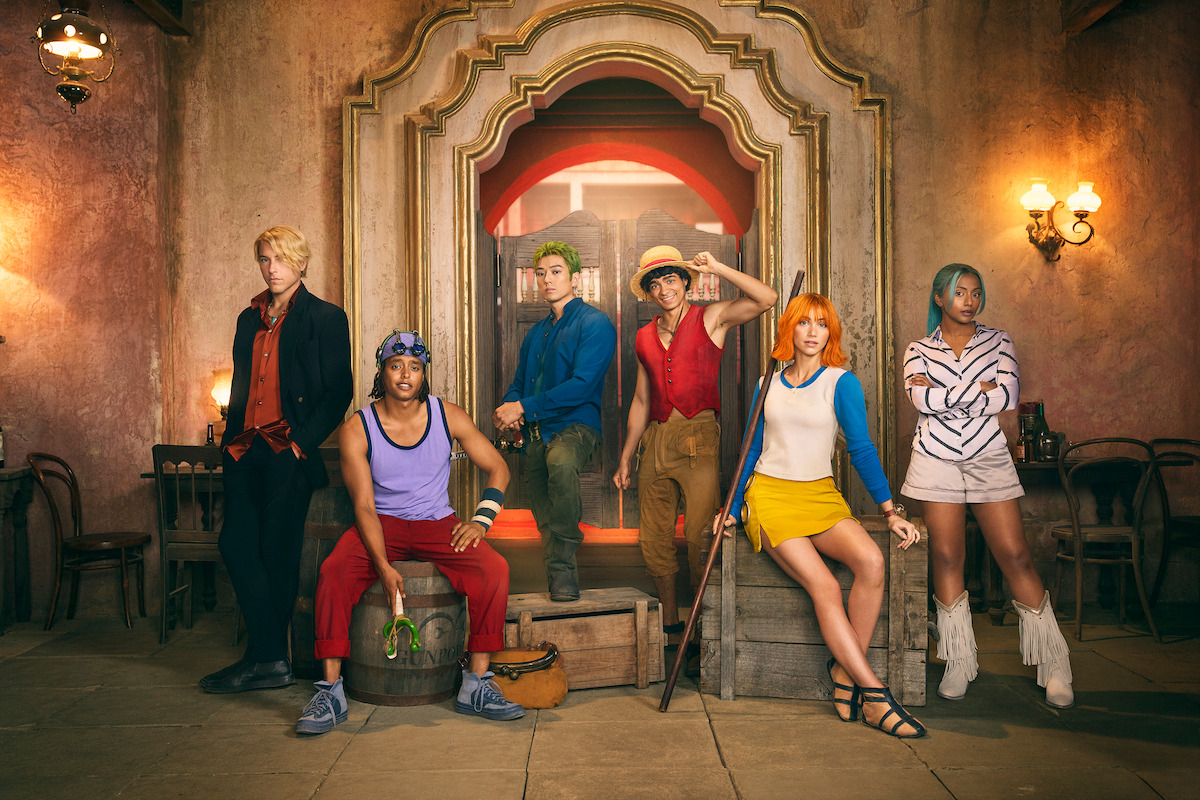 Six people in colorful costumes pose in a vintage, ornate room with old-fashioned lighting, wooden chairs, and stone floor, evoking a fantasy or adventure theme.