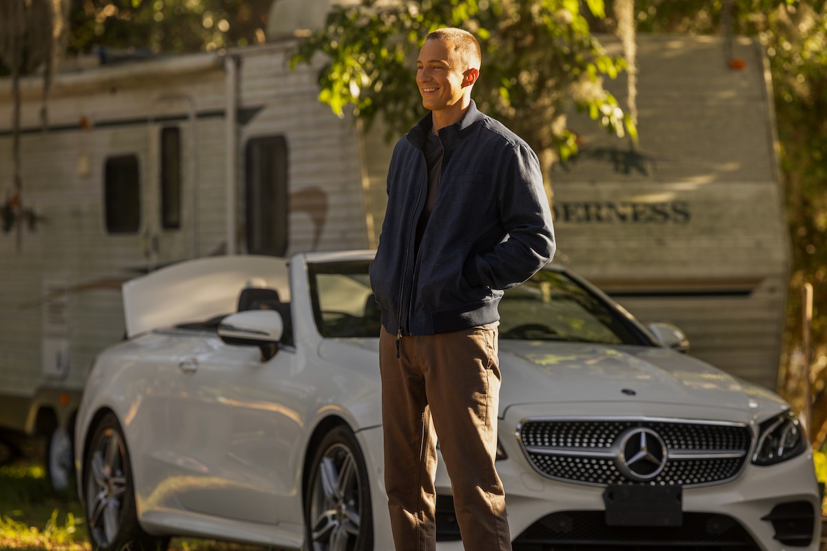 Man standing in sunlight next to a white convertible Mercedes-Benz with a large RV and trees in the background.