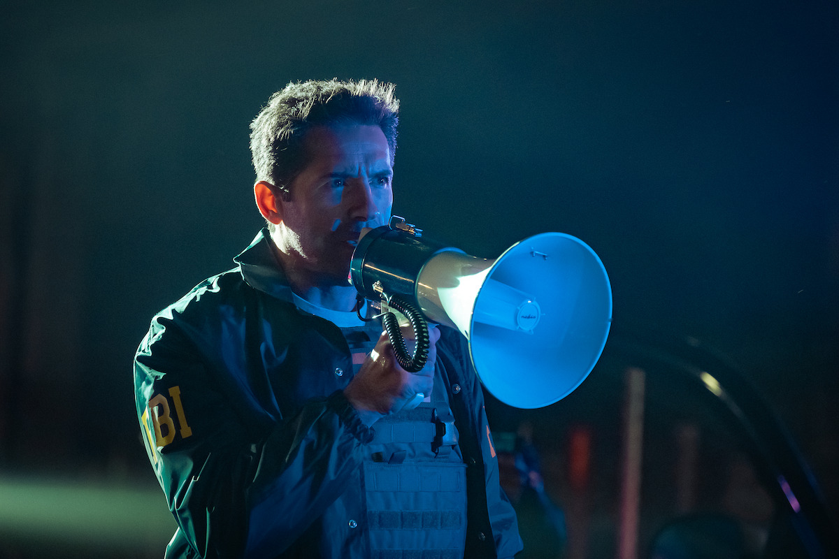 FBI agent speaking into a megaphone at night, wearing a tactical vest and jacket, with dramatic lighting and a blurred dark background, possibly during an operation or emergency situation.