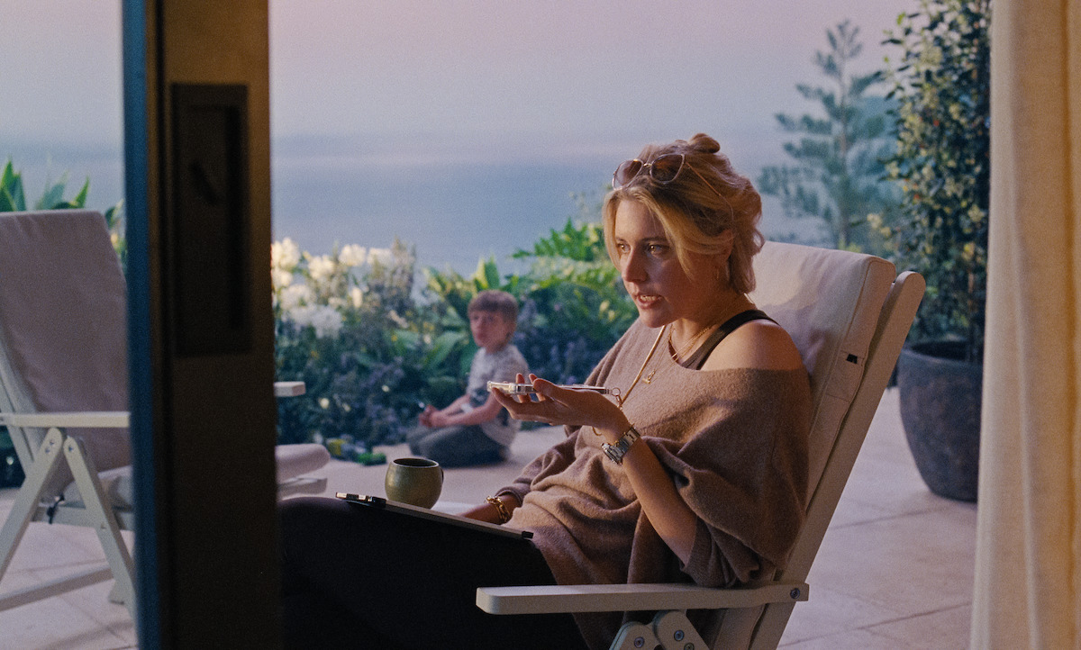 Woman sitting on a patio chair talking, with a mug and notebook, overlooking a garden and sea at sunset; a child is playing in the background, creating a relaxed and introspective mood.