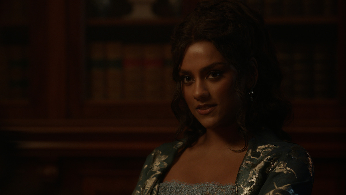 A woman with dark hair in an elegant blue dress sits in a dimly lit library, with shelves of old books in the background, creating a historical and sophisticated atmosphere.