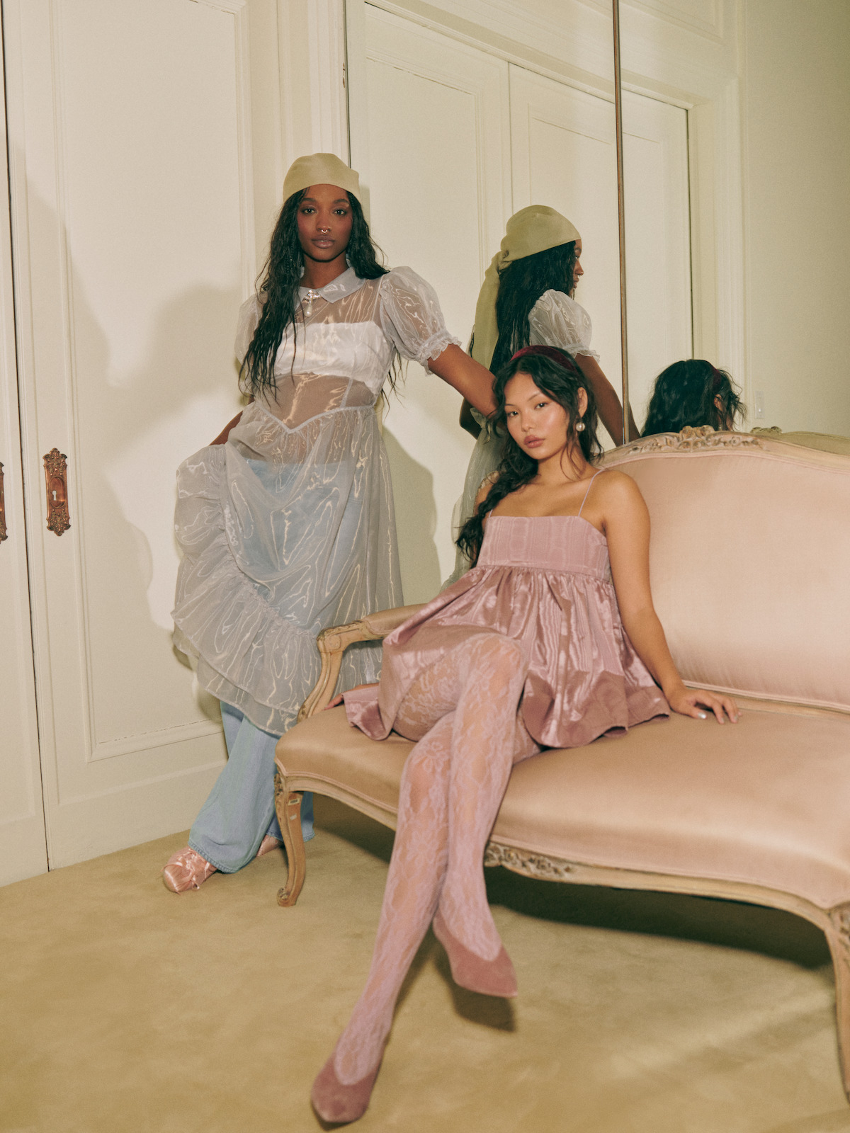 Two women in fashionable sheer and pastel dresses pose in an elegant, vintage-style room with a mirror and ornate furniture, one standing and one seated on a cream sofa.