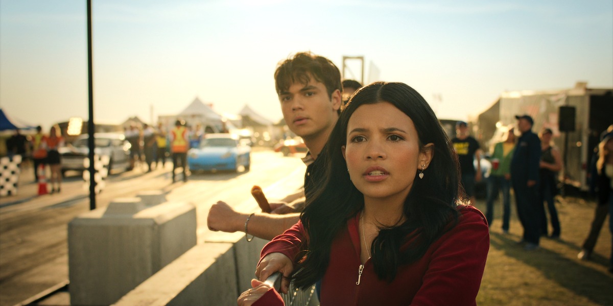 Two young people stand at a racetrack barrier, watching a car race with concern as others look on in the background; the scene is outdoors during golden hour with tents, barriers, and people adding to the vibrant setting.