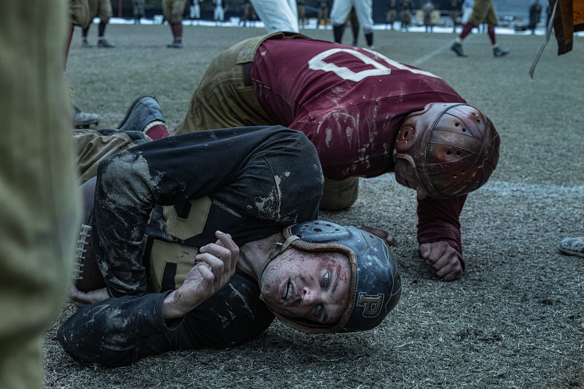 Two vintage football players wearing old-style helmets and uniforms lie on a muddy field after a tackle, both looking exhausted; blurred players and spectators are seen in the background during a gritty, historical game.