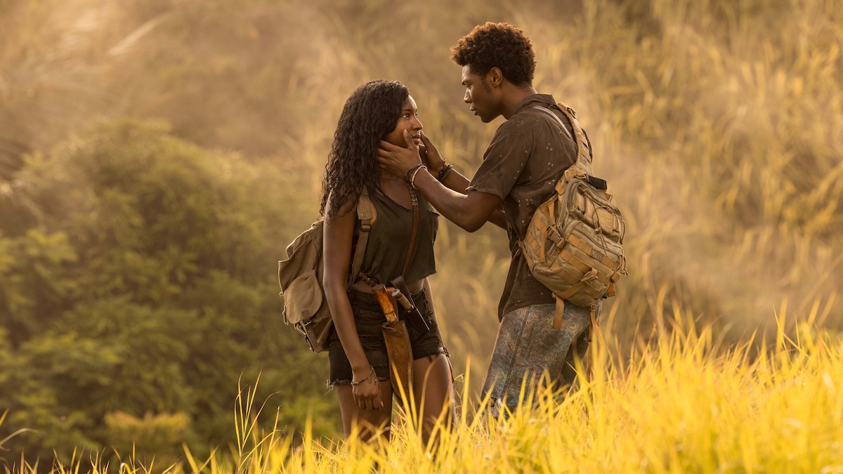 Two young people with backpacks stand in tall grass, one gently holding the other's face, surrounded by a sunlit forest or jungle, suggesting a moment of emotion or connection during an outdoor adventure.