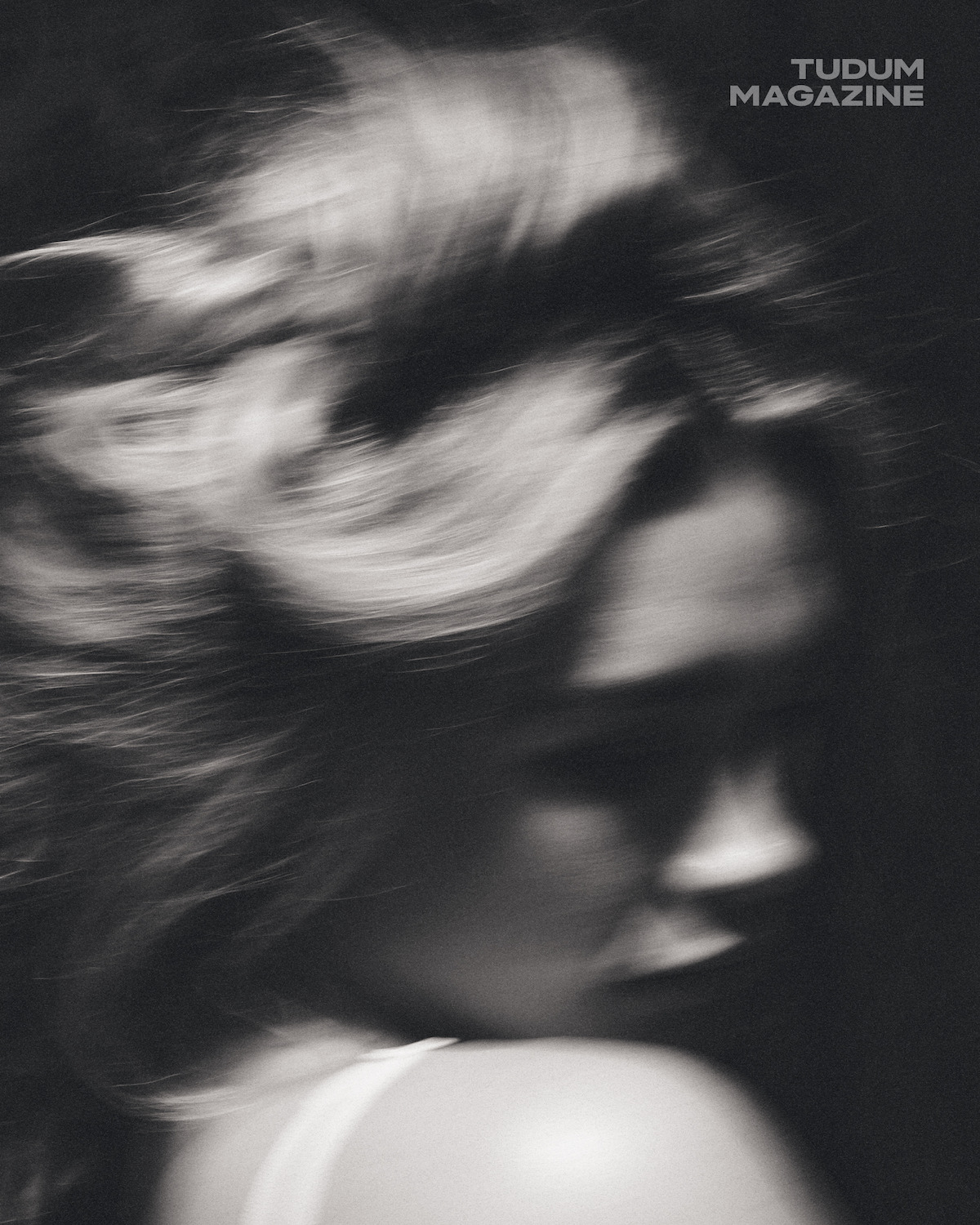 Blurry black-and-white photo of a person with long hair turned away, soft lighting, moody atmosphere; "TUDUM MAGAZINE" text in the upper right corner.