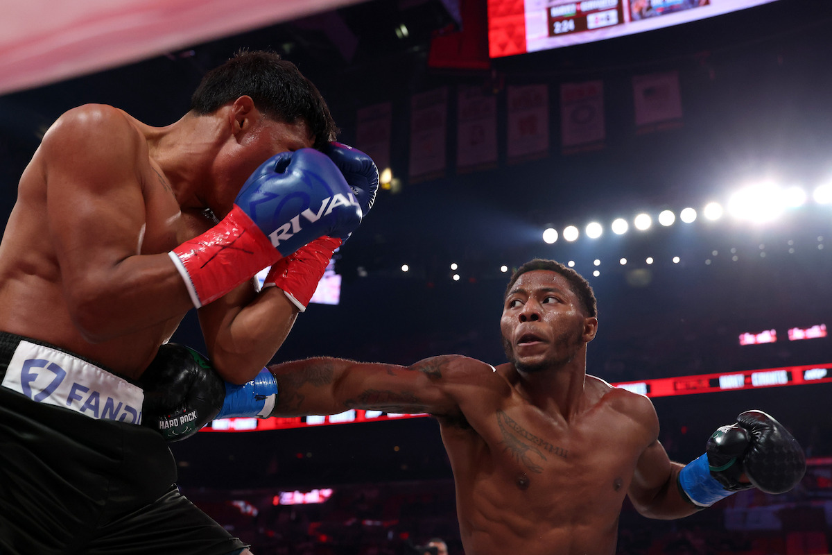 Kevin Cervantes and Jahmal Harvey in the ring during a match, one defending with gloves up against the ropes, the other throwing a punch, with bright arena lights and a crowd in the background.