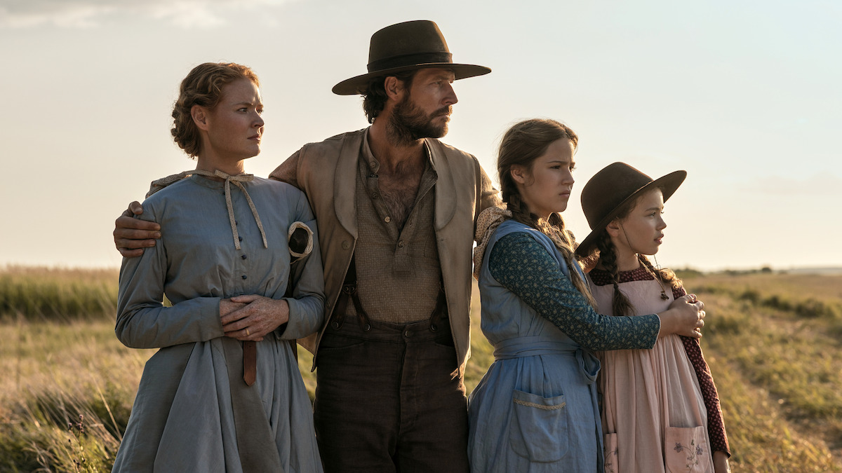 A man and three girls in old-fashioned clothing stand close together in a grassy field, looking intently into the distance under an open sky, evoking a historical or pioneer setting.