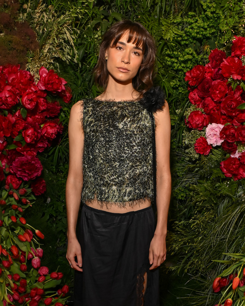 Woman in a textured sleeveless top and black skirt stands in front of a lush floral backdrop with red and pink flowers and greenery, posing with a neutral expression.
