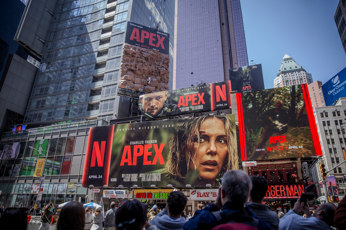 Crowd in Times Square with large digital billboards advertising Netflix’s “Apex” movie, featuring actors’ faces and dramatic visuals on tall buildings in an urban city environment during daylight.