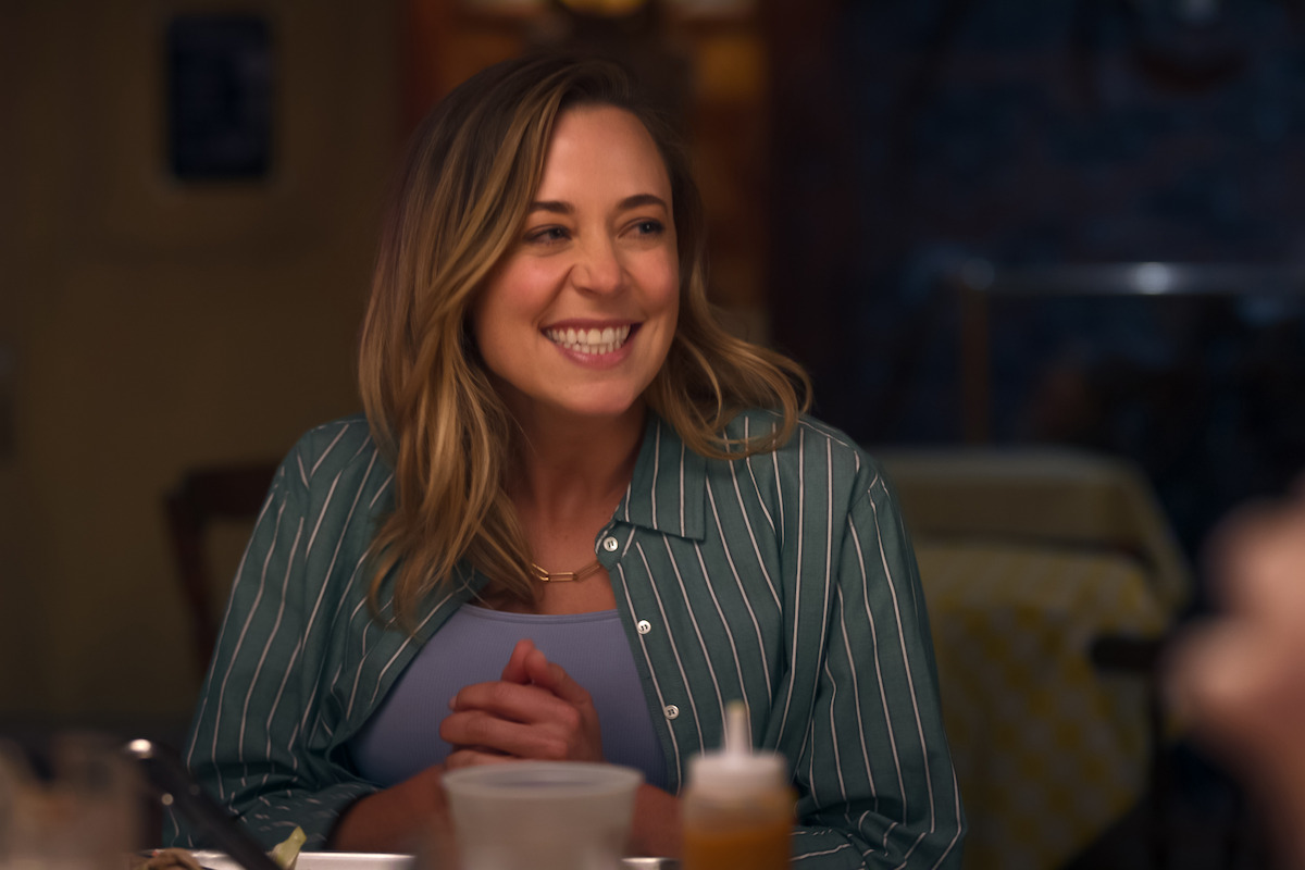 A woman with long hair and a striped shirt smiles while sitting at a dining table in a cozy, warmly lit restaurant or cafe. Food and drinks are visible on the table around her.