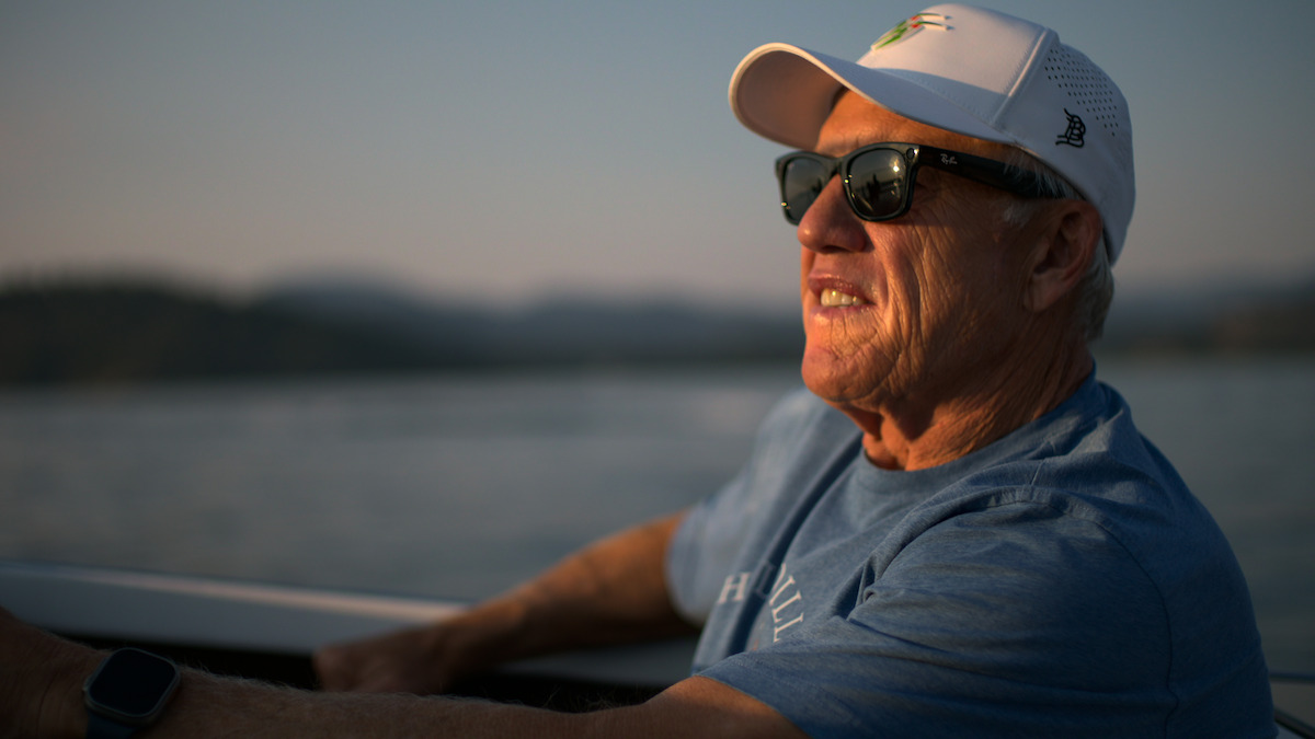 John Elway wearing sunglasses, a white cap, and a blue shirt sits outdoors on a boat, smiling, with soft evening light and blurred water and hills in the background.