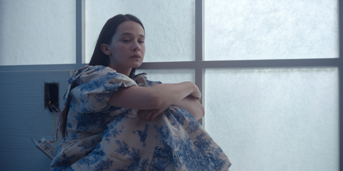 Young woman in a floral dress sits on the floor by a frosted window, arms around knees, looking thoughtful in a softly lit, minimalist room with exposed wires on the wall.