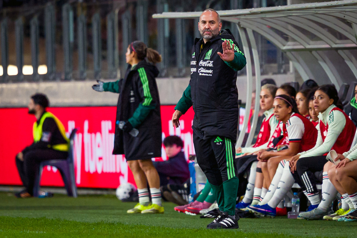 Soccer coach gesturing on the sideline during a match, with players and staff seated on the team bench in a stadium environment, bright field lights and advertisement boards visible in the background.