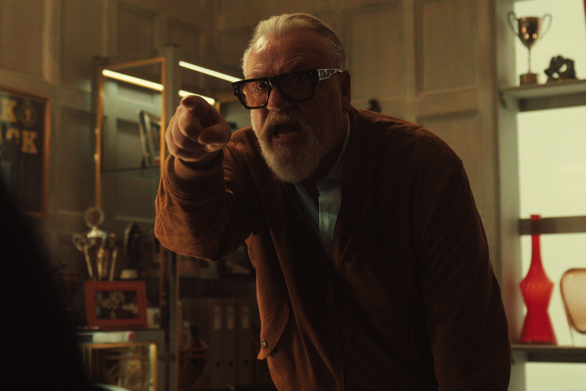 Older man with white hair and beard, wearing glasses and a brown jacket, angrily pointing in a modern office with trophies, shelving, and decorative items in the background.