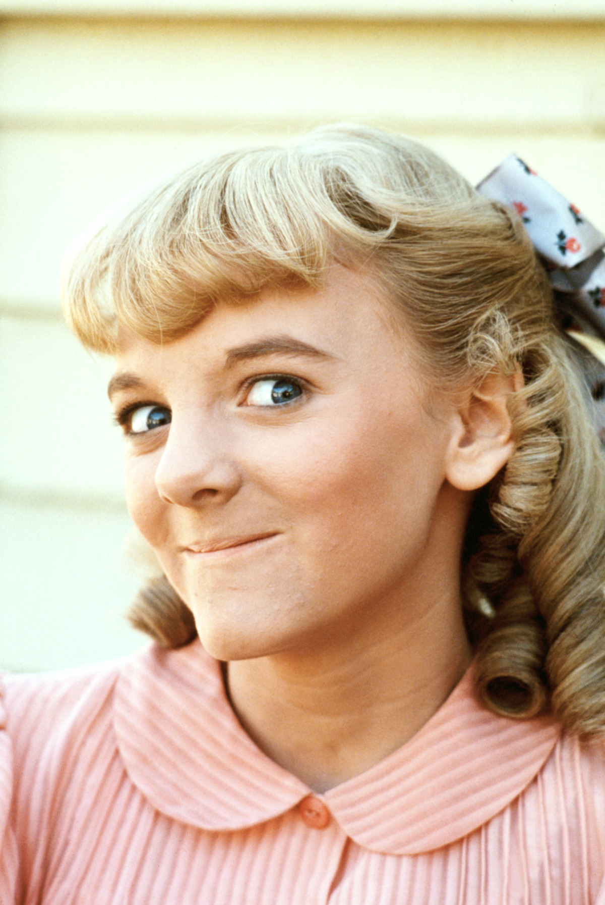 Young girl with blonde curled hair, a bow, and a pink collared dress smiles mischievously at the camera, standing in front of a light-colored wall in a brightly lit setting.