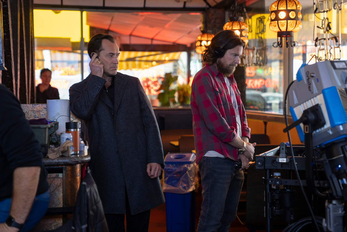 Two men stand in a diner filled with film equipment and lighting; one wears a dark coat, the other a plaid shirt, both appear focused, suggesting a movie or TV production setting with a casual, behind-the-scenes atmosphere.