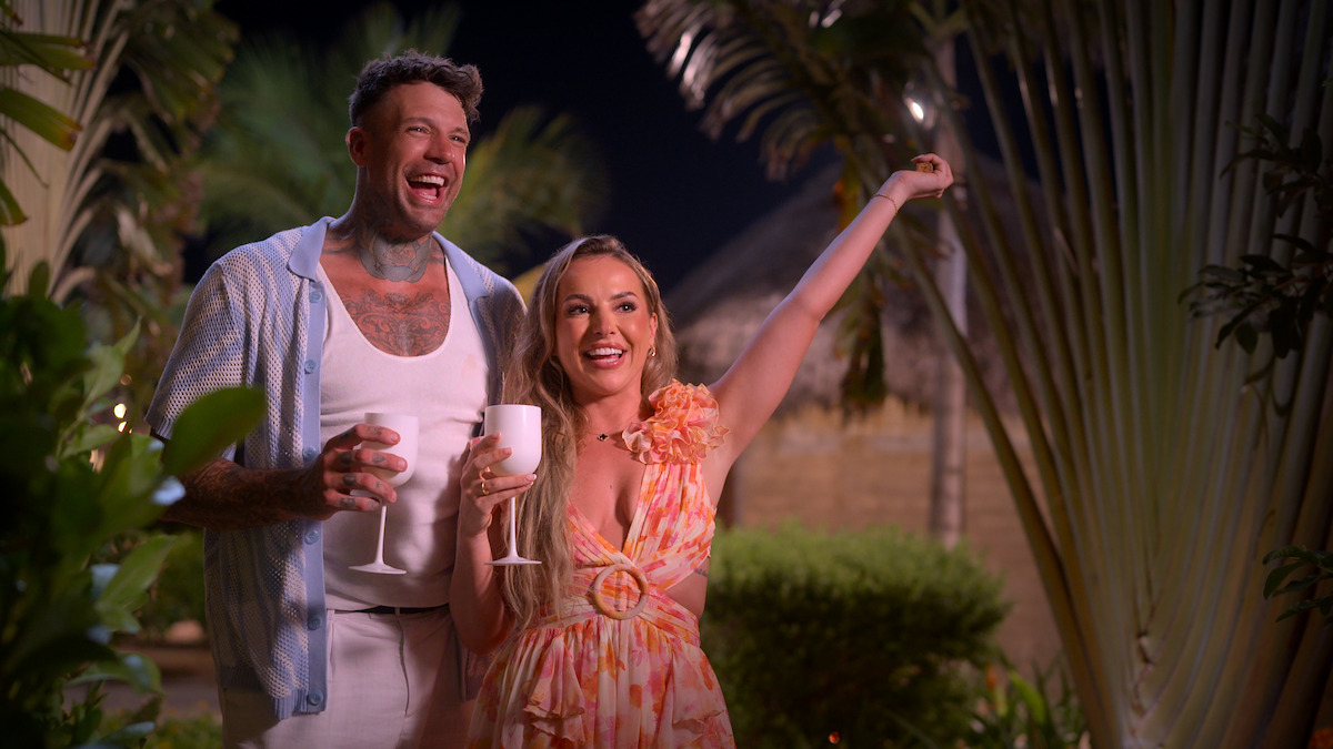 A smiling man and woman holding white drink glasses stand together outdoors at night, surrounded by tropical plants and palm trees, with thatched-roof huts in the background. The woman raises her arm joyfully.