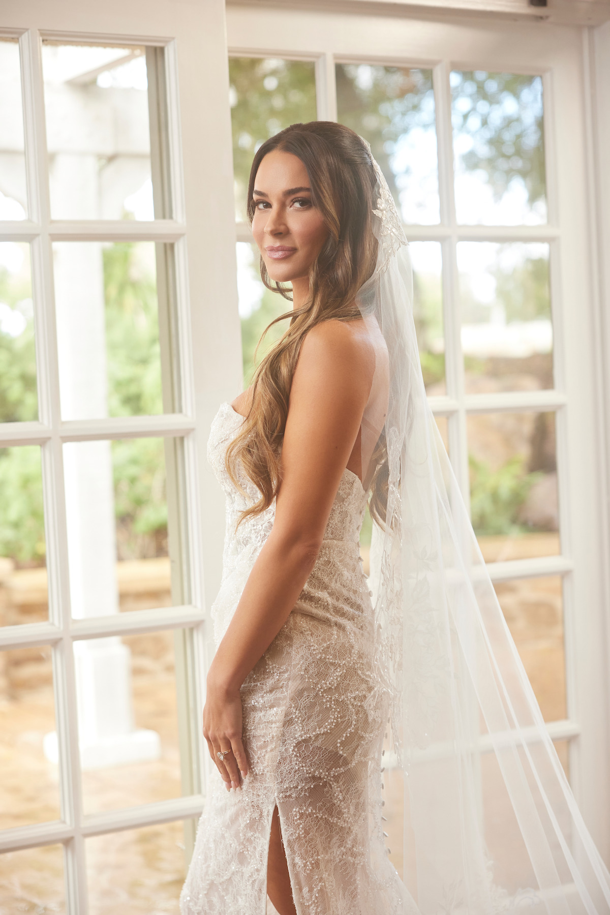 Bride in lace wedding gown and veil stands indoors by large glass doors, with soft natural light and outdoor greenery visible through the windows, creating an elegant and serene atmosphere.