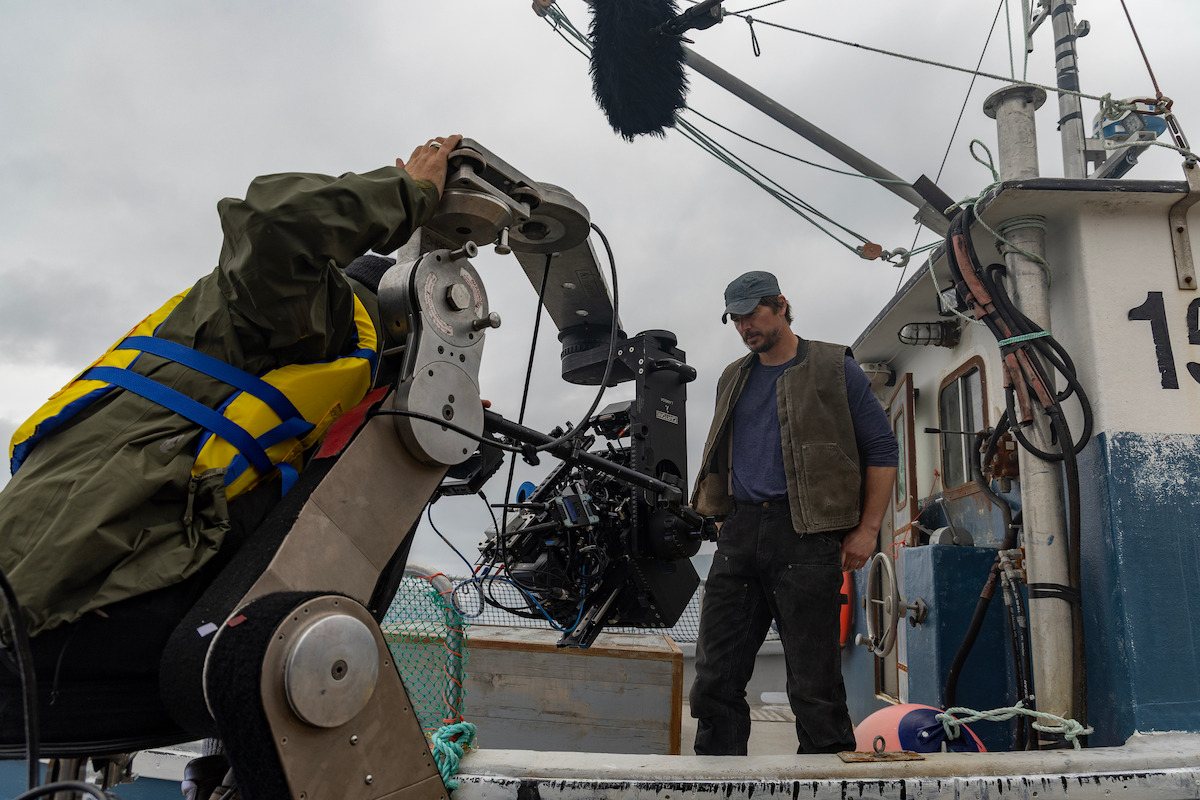 Film crew shoots a man standing on the deck of a fishing boat, cloudy sky above, with camera and boom microphone equipment visible, capturing a maritime scene in an outdoor working environment.