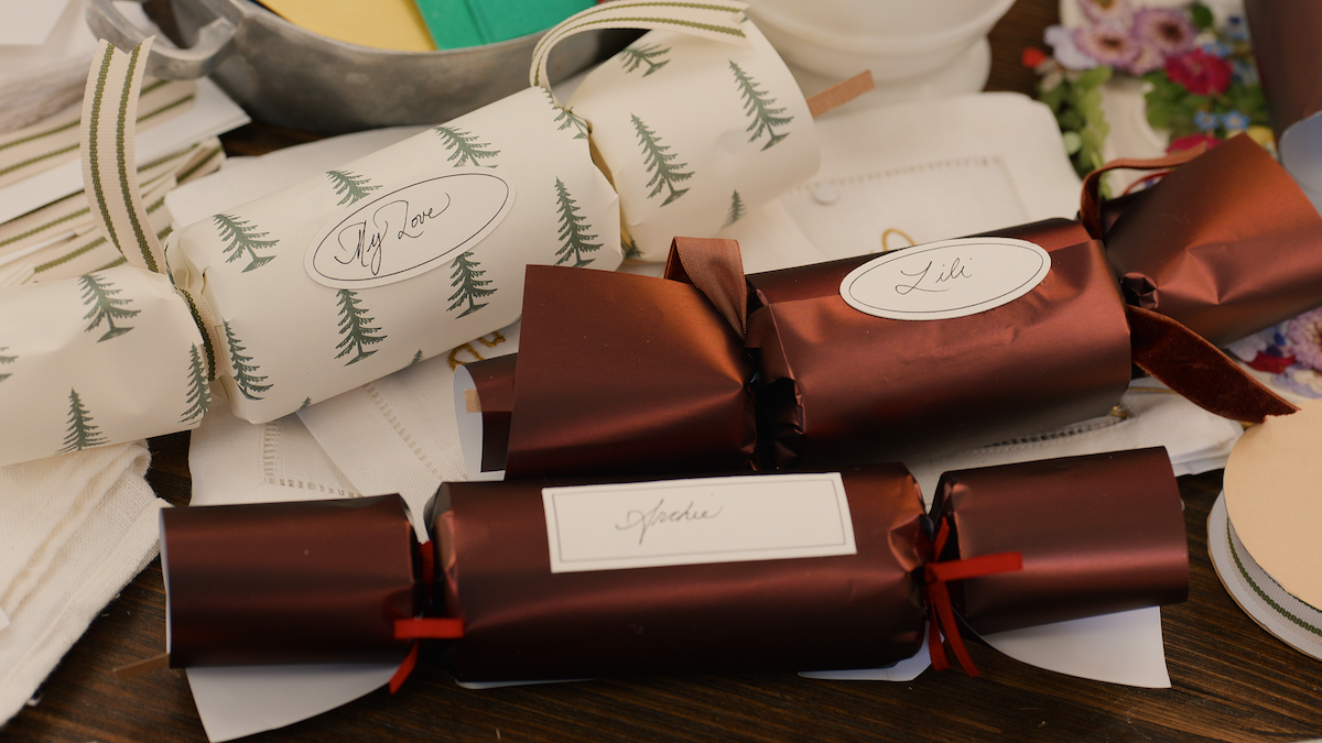 Three festive Christmas crackers, two maroon and one white with green trees, labeled with names on a decorated table, surrounded by napkins, flowers, and warm ambient lighting, creating a cozy holiday mood.
