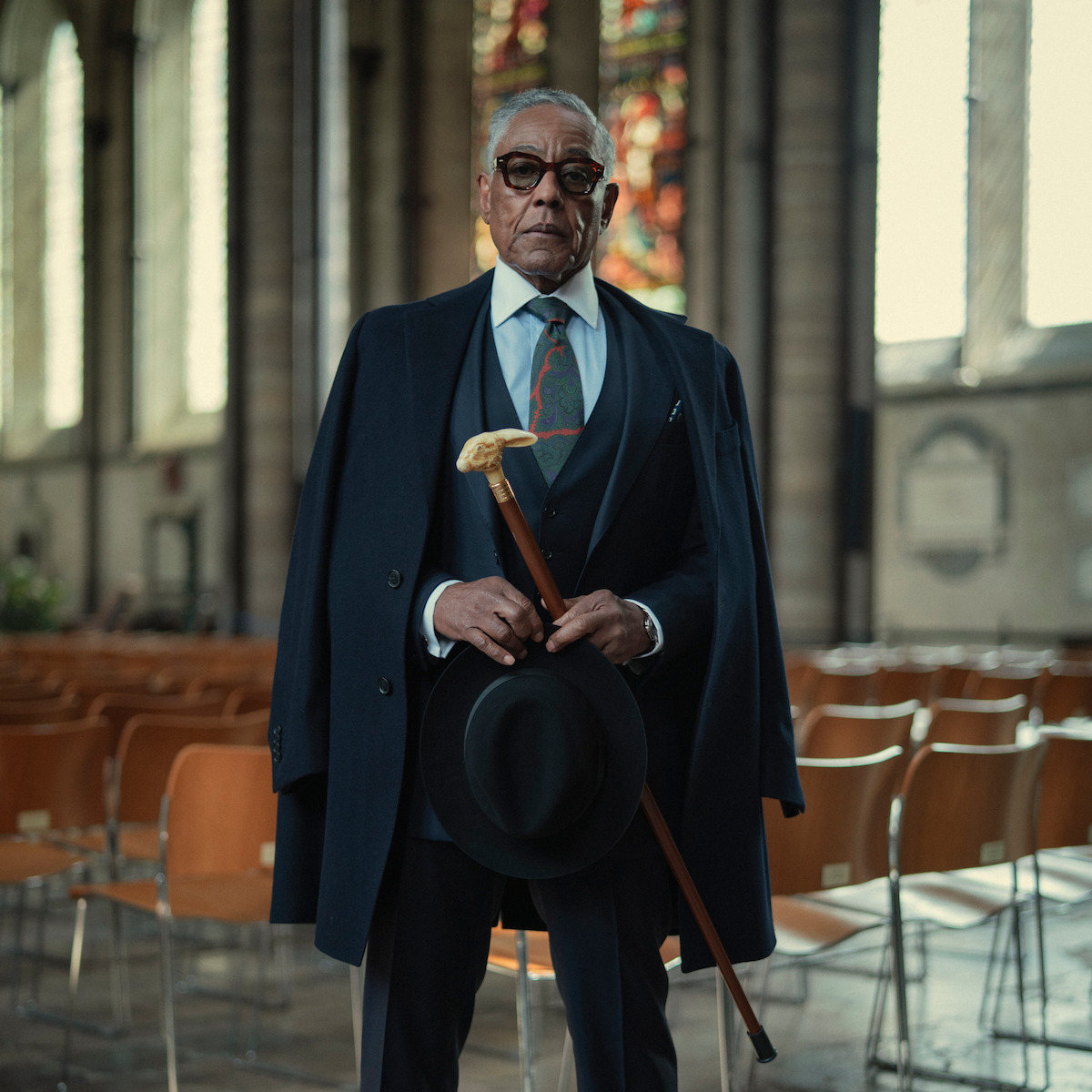 Elderly man in a suit and overcoat stands in an empty, sunlit church holding a cane and a hat, with stained glass windows and wooden chairs in the background.