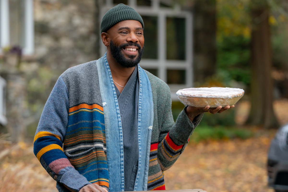 Smiling man outdoors in autumn, wearing a striped cardigan and green beanie, holding a pie in one hand, with a house and trees in the background.