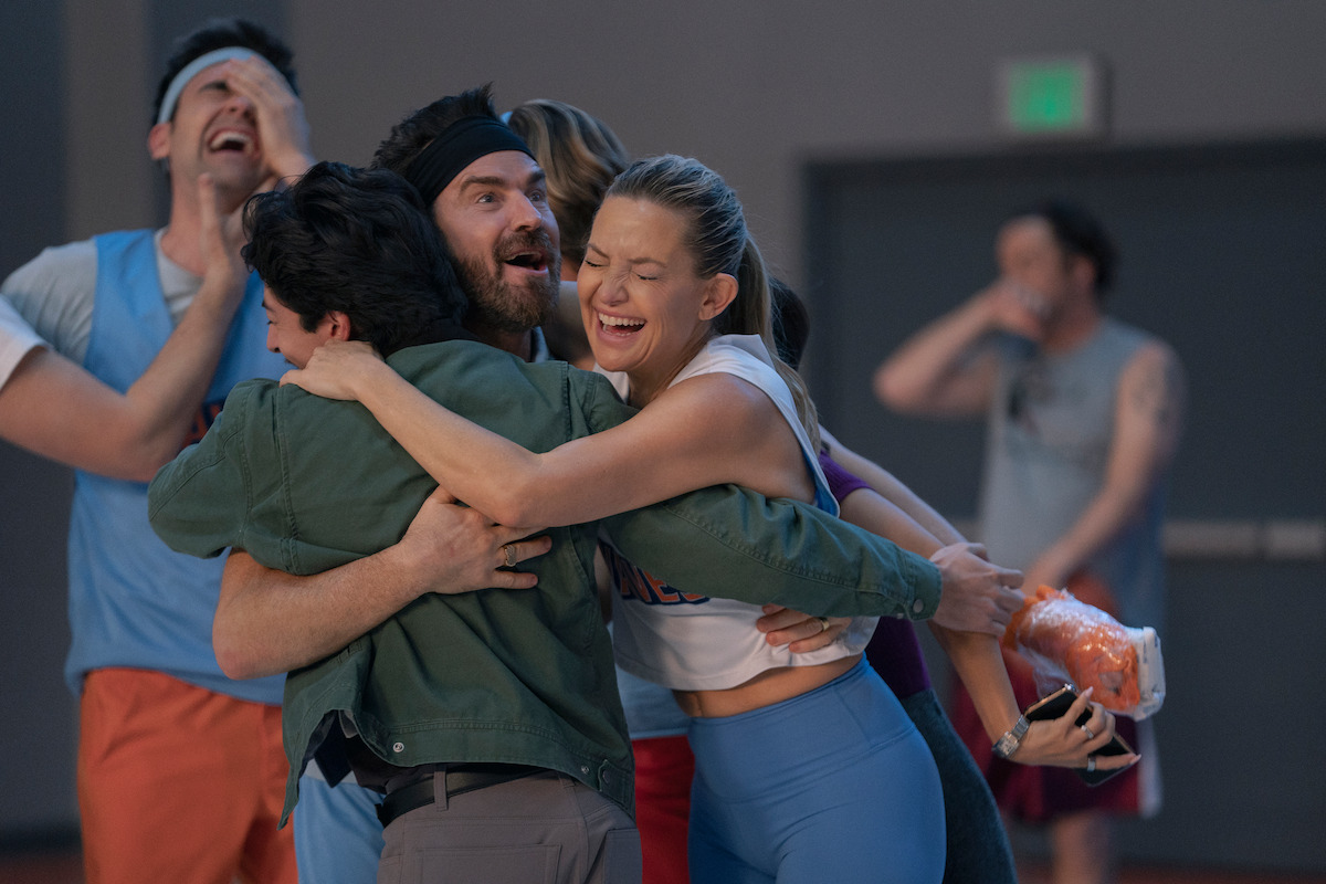 Group of people celebrating and hugging in a gymnasium, with some laughing and one holding a bag of carrots, suggesting a joyful and energetic environment during an indoor sports or recreational activity.