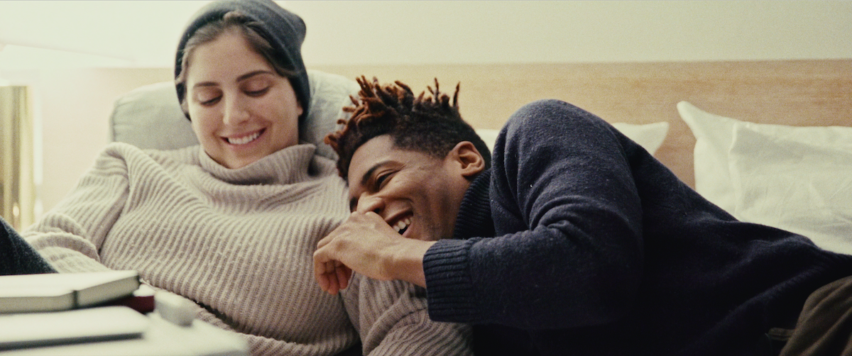 Suleika Jaouad, wearing a cream turtleneck sweater, and Jon Batiste, in a black sweater, smile as they sit in bed.