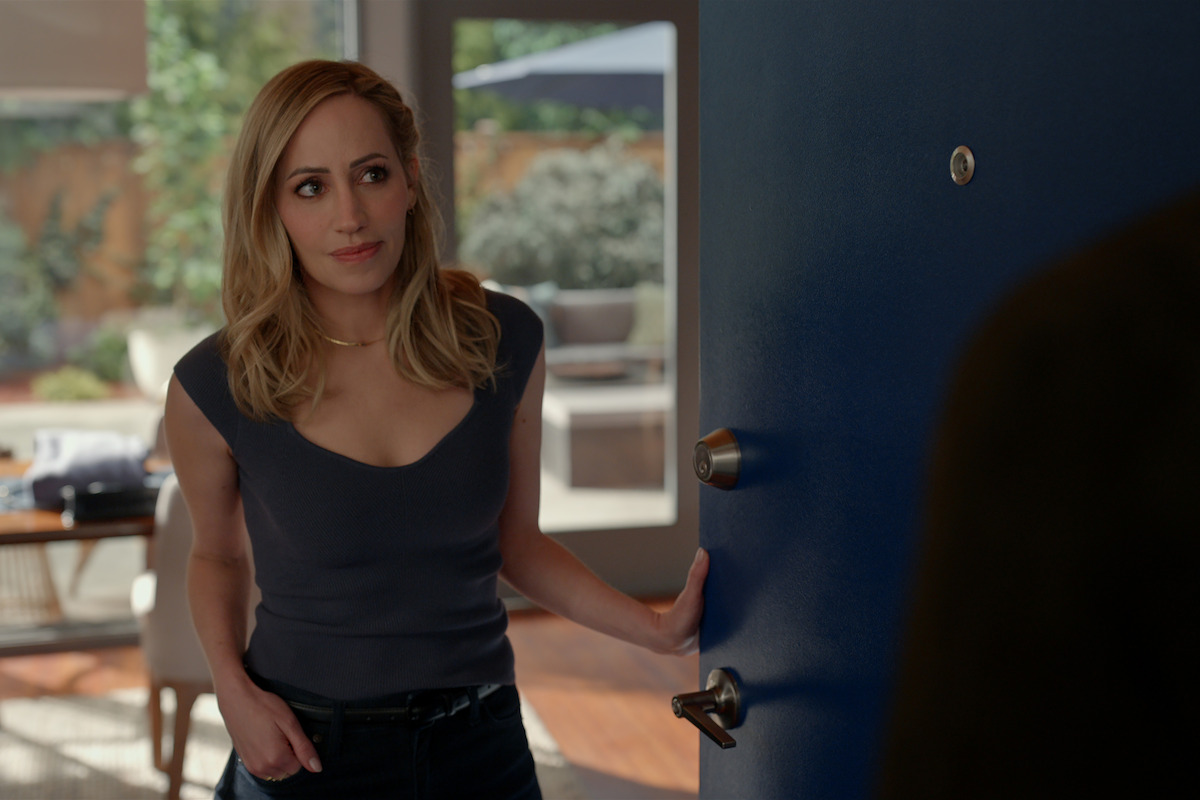 A woman with blonde hair stands at a blue door, looking out, one hand in her pocket in a modern, sunlit home interior with a garden visible through large windows in the background.