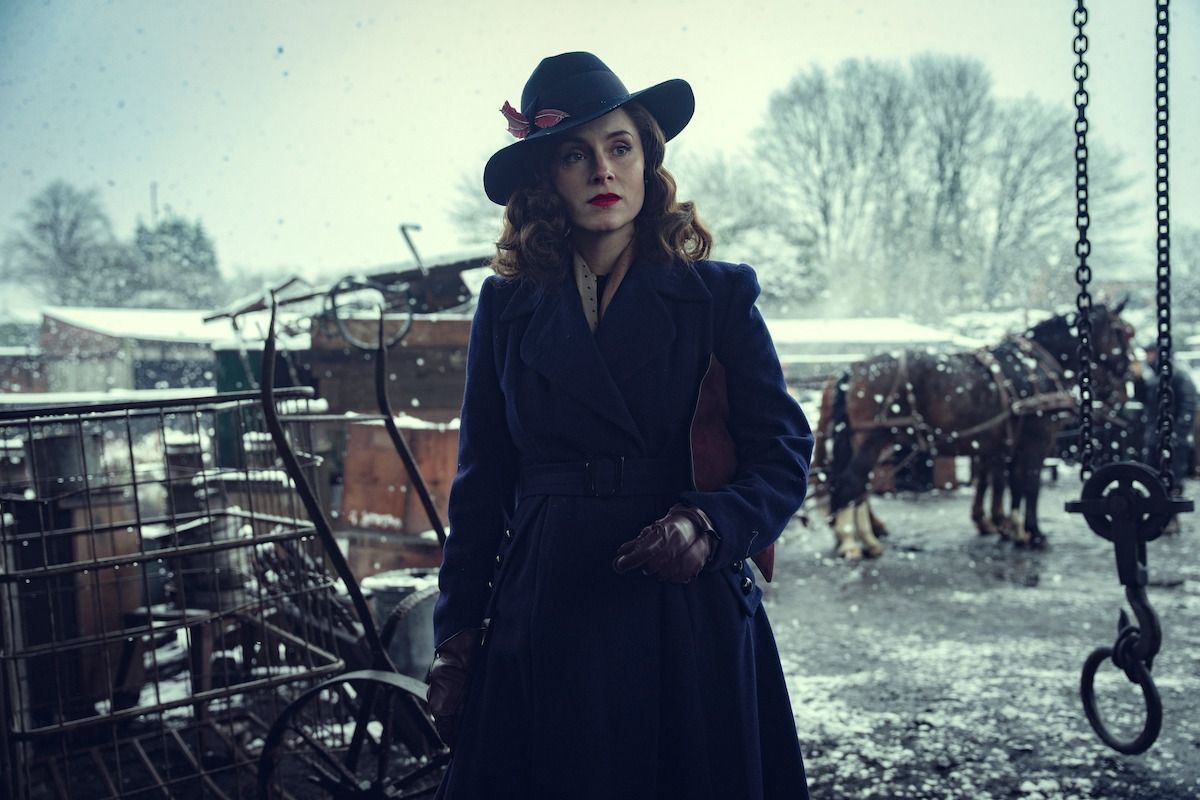 Woman in vintage coat and hat standing outdoors in snowy, wintry setting, with old carts and horses in the background, evoking a historical or period atmosphere.
