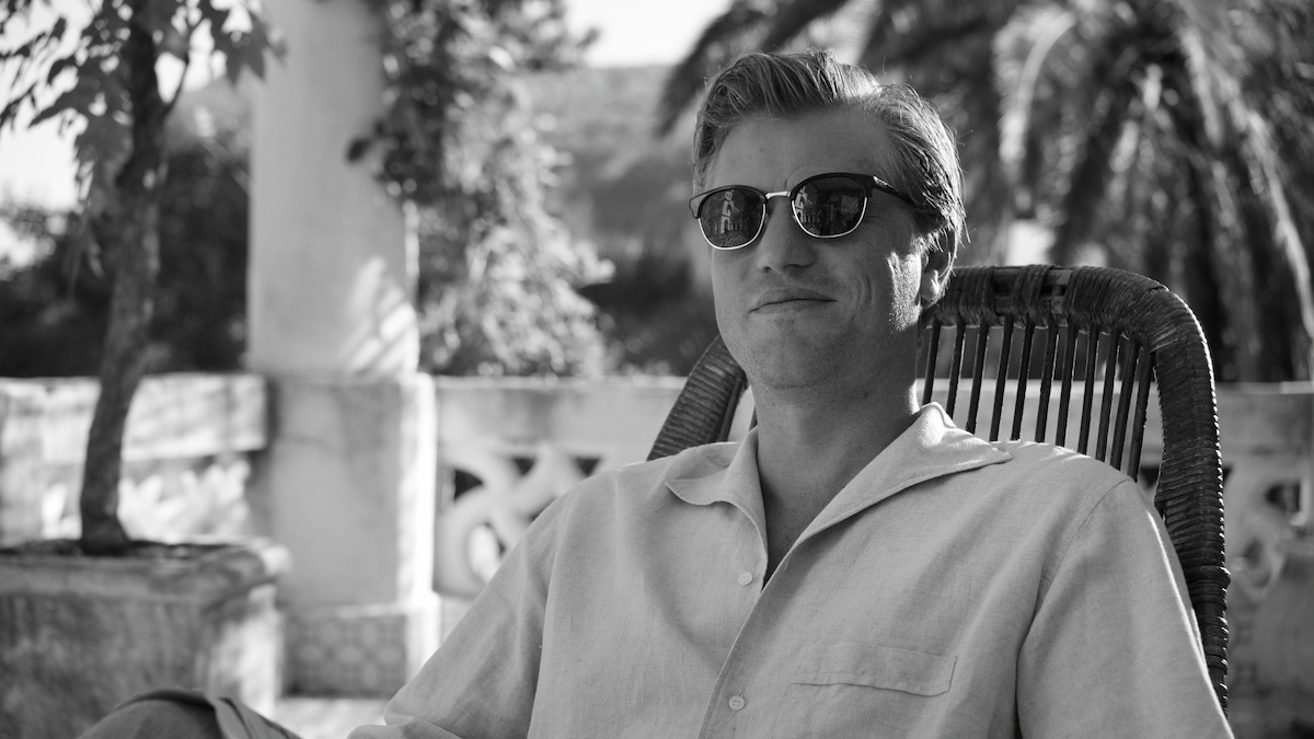 Black-and-white photo of Johnny Flynn as Dickie Greenleaf wearing sunglasses and smiling in ‘Ripley’