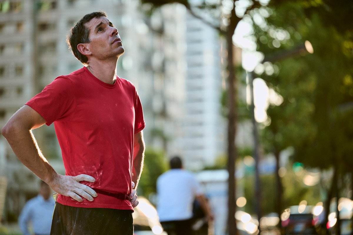 Alex Honnold, con una camisa roja, parado al aire libre en un entorno urbano, mirando hacia arriba con las manos en las caderas, luciendo pensativo o cansado; edificios y árboles visibles al fondo con la luz del sol filtrándose.