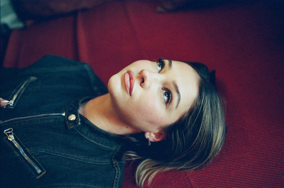 Woman with light hair lying on a red couch, looking upwards in a relaxed indoor setting, wearing a dark denim jacket with gold zippers. Soft lighting and shallow depth of field create a calm and intimate mood.