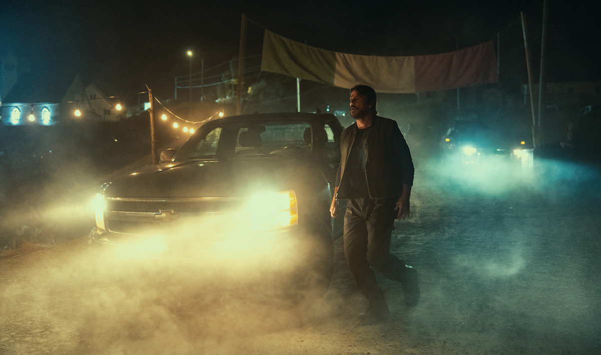 A man walks in front of a truck with headlights on, surrounded by smoke at night. String lights, a colorful banner, and another vehicle are visible in the background, creating a dramatic and intense atmosphere.