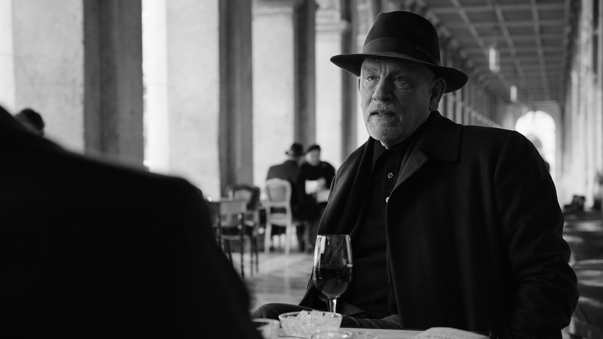 John Malkovich as John Malkovich sits at an outdoor cafe table in ‘Ripley’