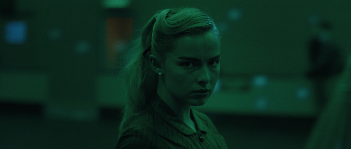 A woman with blonde hair and serious expression stands in a dimly lit, green-tinted indoor environment, with blurred figures and abstract lights in the background, creating a tense and mysterious mood.