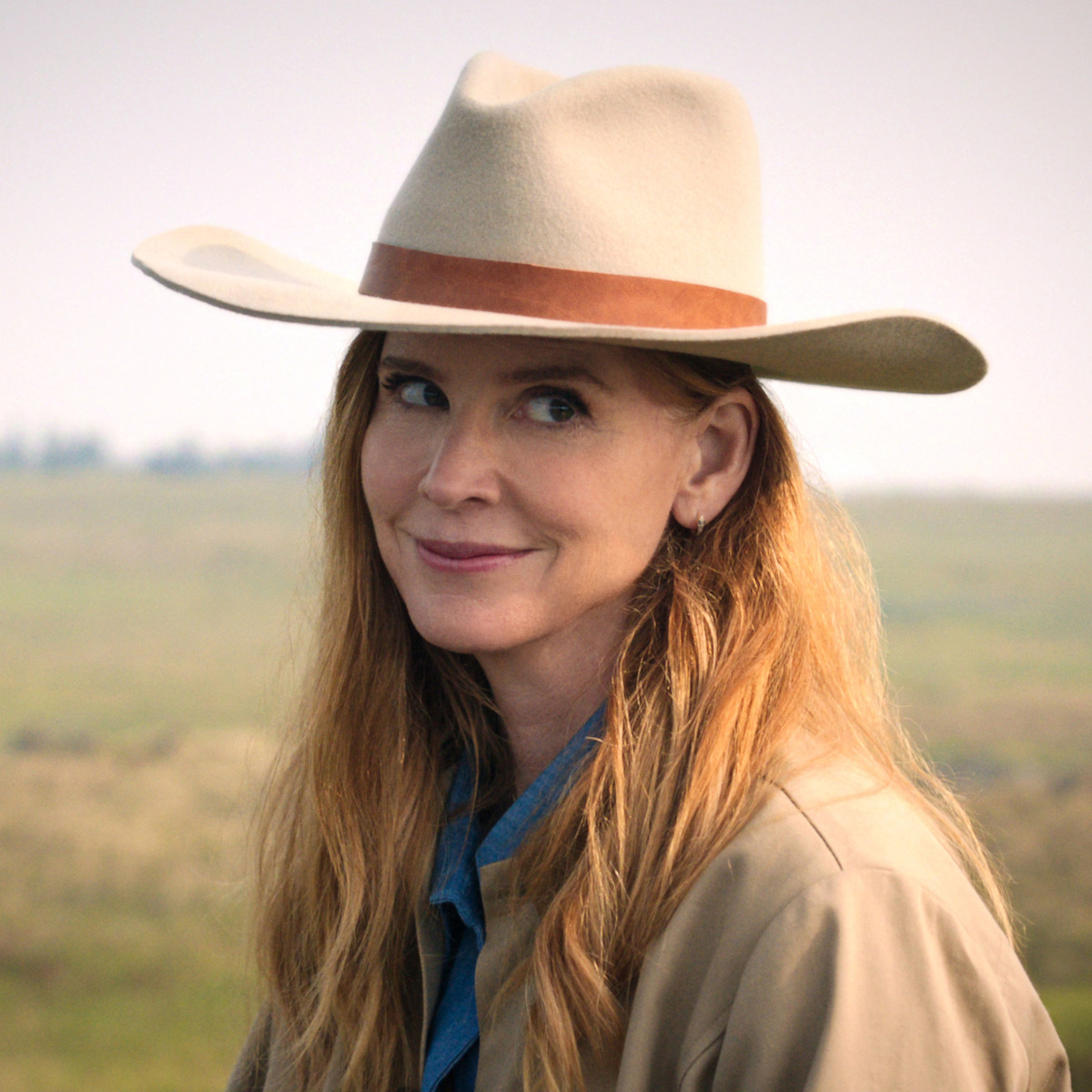 Smiling woman with long hair and a tan cowboy hat outdoors, wearing a tan coat and denim shirt, standing in an open grassy field with a blurred sky and horizon in the background.