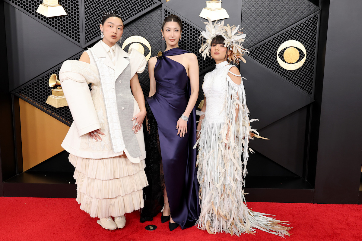 Three people in creative, high-fashion outfits pose on a red carpet at an awards event, standing in front of a black and gold geometric backdrop with spotlights and music award logos.