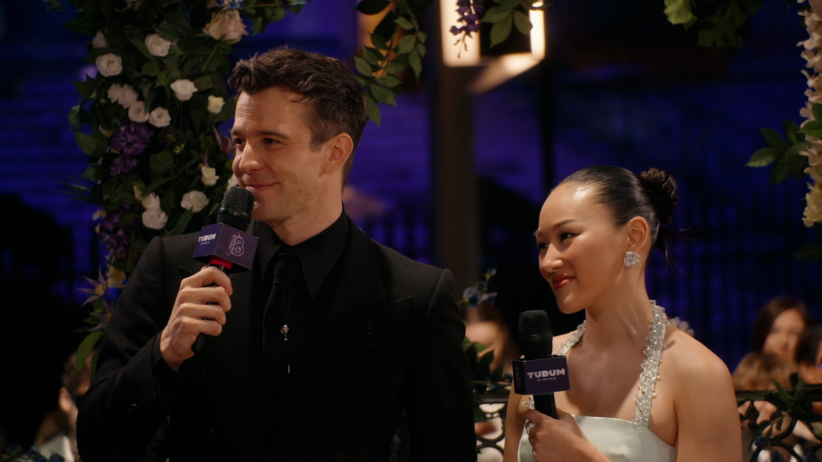 Luke Thompson in a black suit and Yerin Ha in a white dress hold microphones, smiling and speaking at an elegant event decorated with flowers and soft lighting, surrounded by an audience in formal attire.