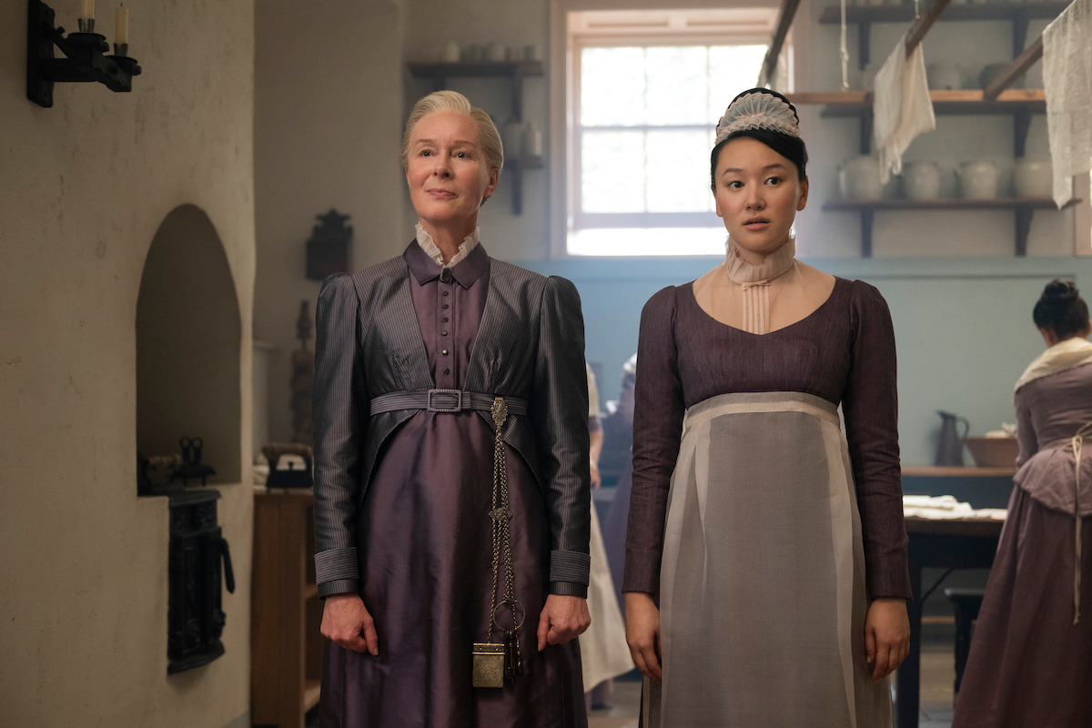 Two women in period costumes stand side by side in a historical kitchen, lit by a window. The older woman smiles slightly, the younger appears serious. Shelves and kitchenware fill the background.