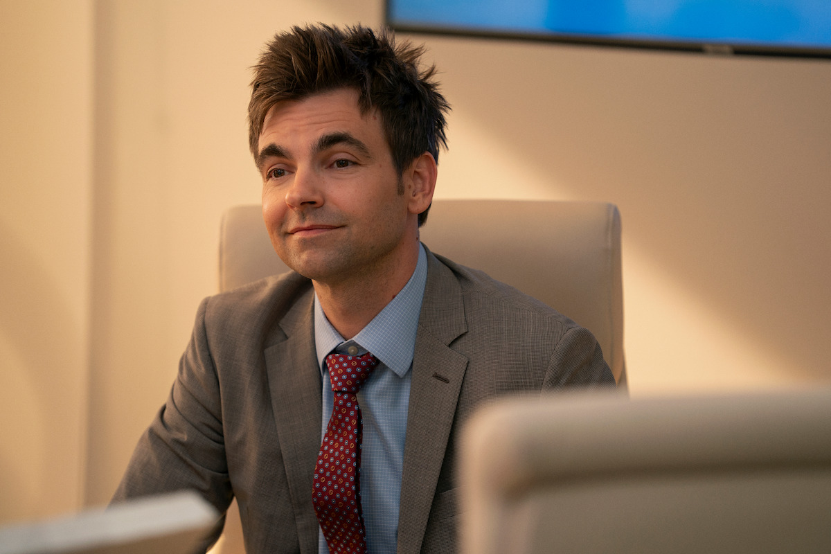 A man in a gray suit and red tie sits at a conference table in an office setting, appearing to be in a meeting with a neutral background and a blurred screen behind him.