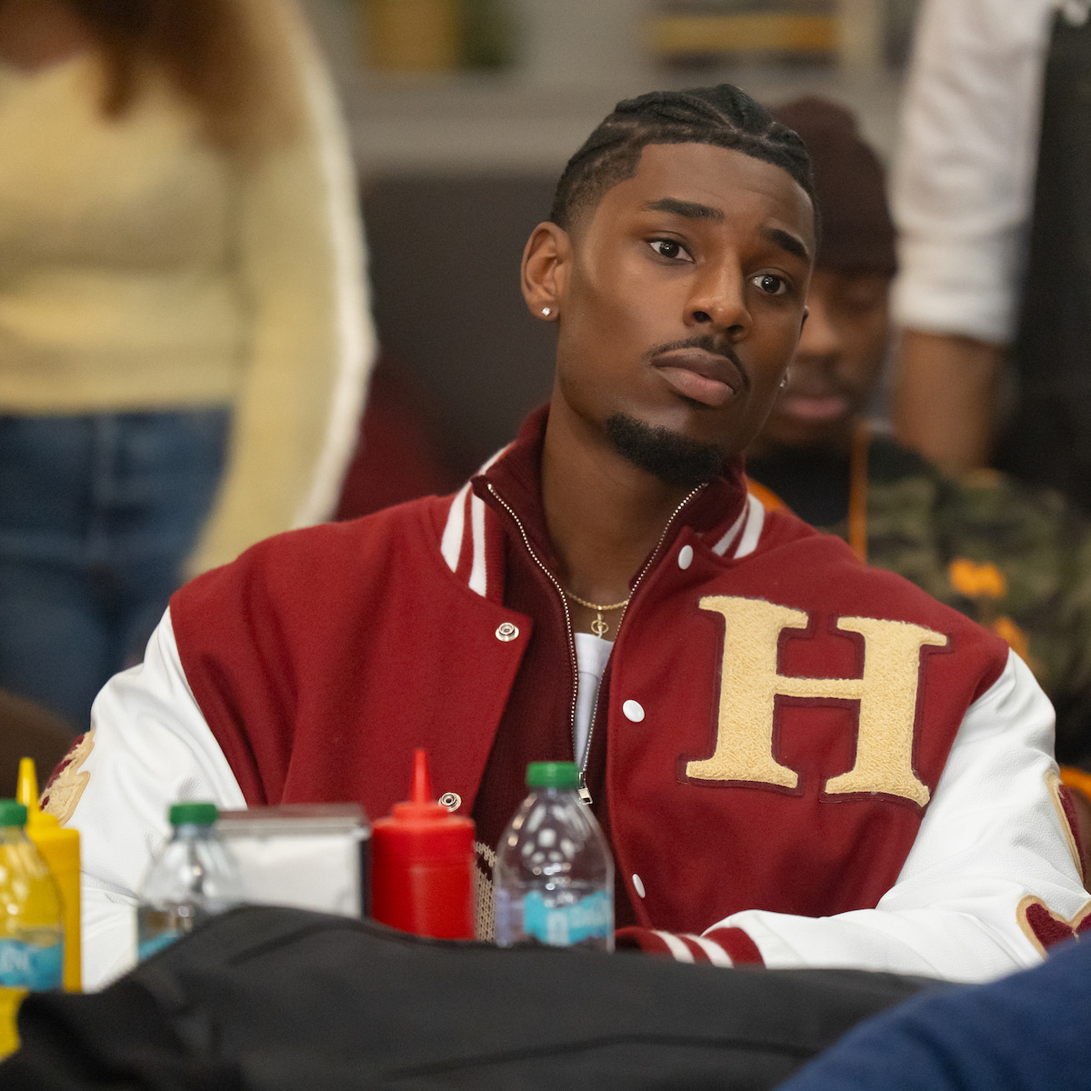 Joshua Suiter as Andre Tilsdale in a red letterman jacket with an "H," sitting indoors at a table with bottles and condiments, looking thoughtful, blurred people and casual setting in background.