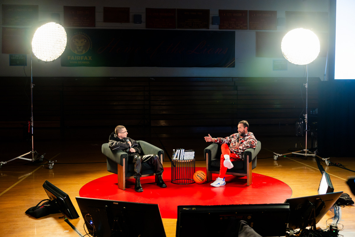 Two people have a conversation on stage in a brightly lit gymnasium, seated in armchairs on a red carpet, surrounded by studio lights, monitors, and a basketball, creating a professional yet relaxed atmosphere.