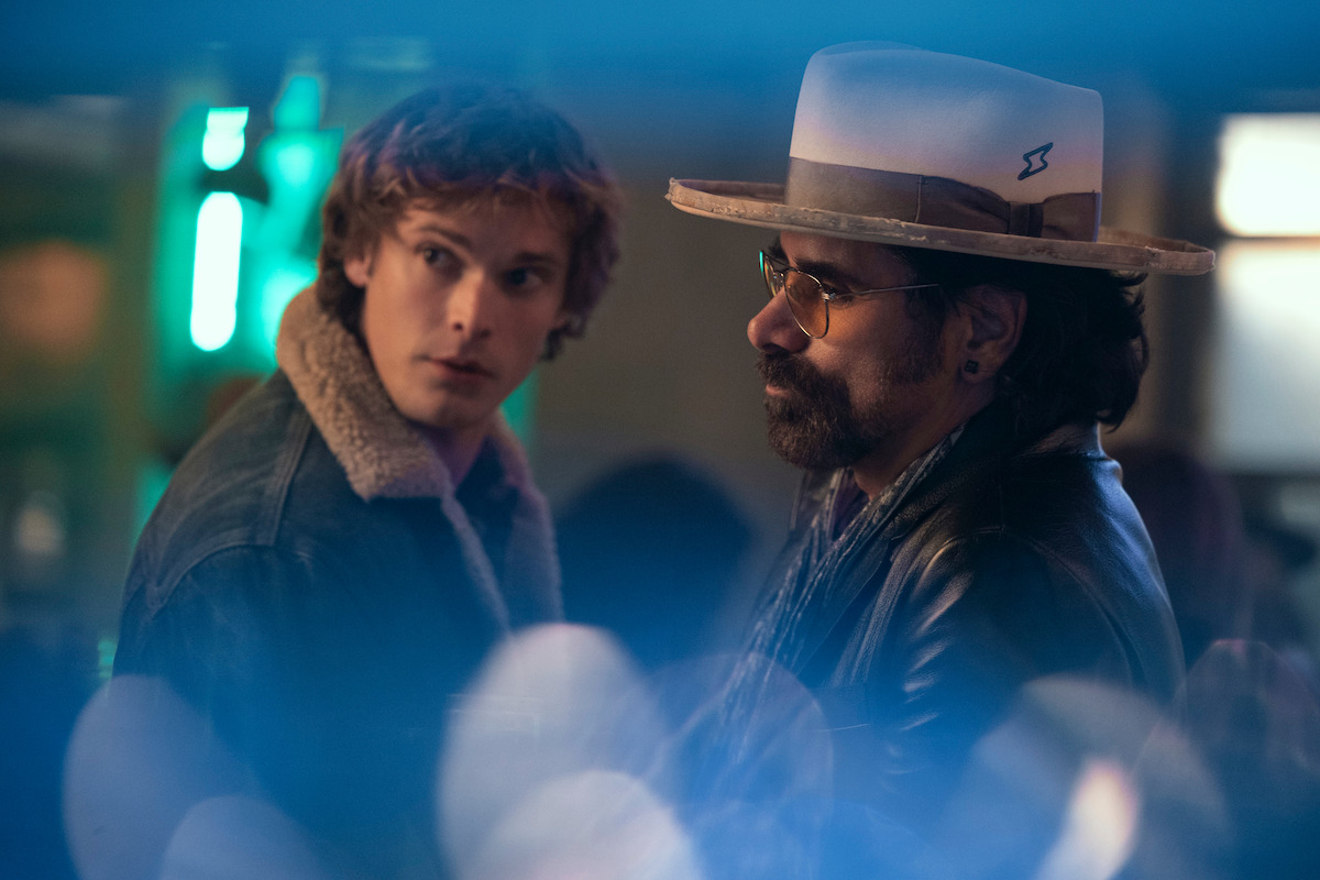 Two men in a dimly lit indoor setting, one in a jean jacket with a fur collar looking back, the other with a hat and glasses facing forward, colorful lights blurred in the foreground.