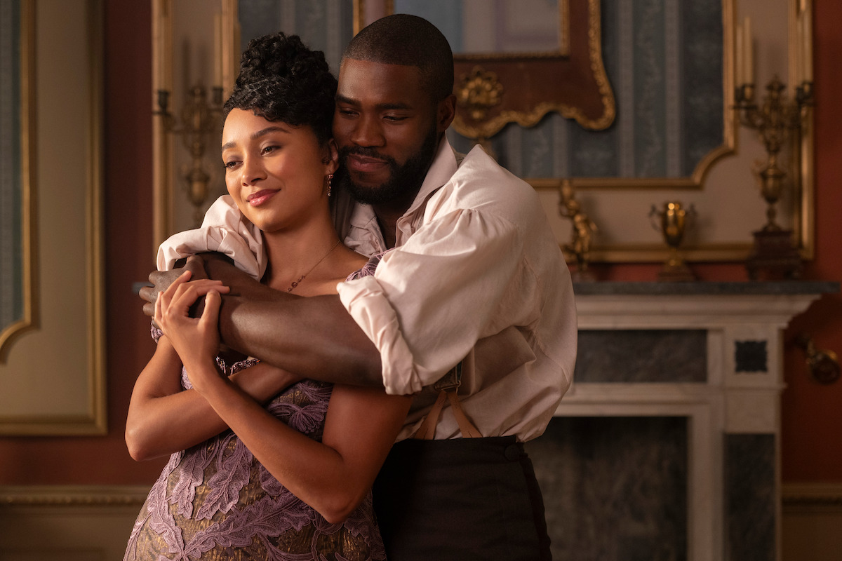 A couple dressed in historical attire embrace in an elegant, ornate room with gold accents, a marble fireplace, candelabras, and a framed mirror in the background.
