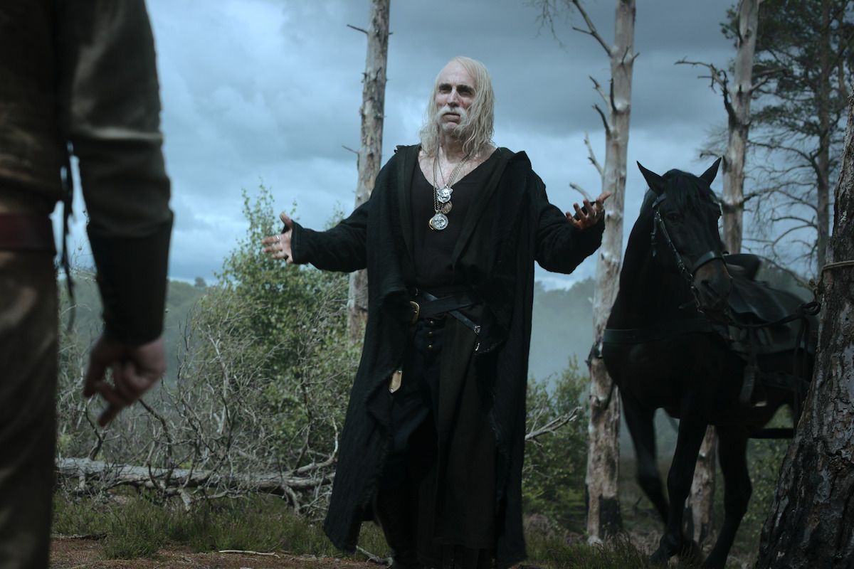 An elderly man with long white hair and black robes stands in a misty forest with bare trees, arms outstretched, facing another figure; a black horse stands in the background, creating a tense and mystical mood.
